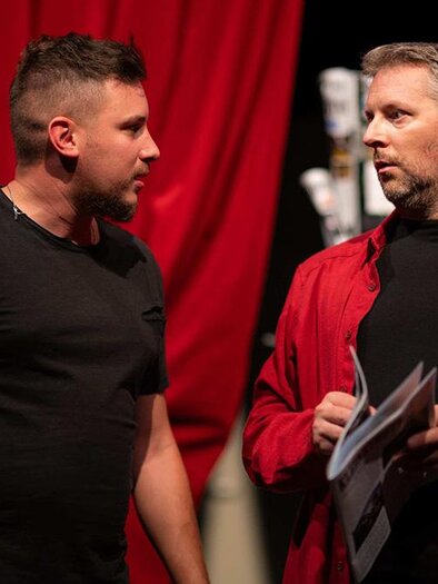 Two men are standing on a stage with red curtains in the background. One of them is holding a sheet of paper while they converse. | © Bild: Felix Kainrath-Reumayer