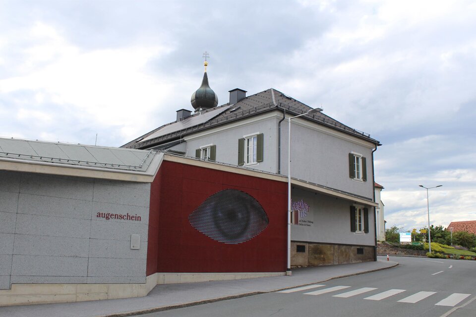 Ein modernes Gebäude mit einem auffälligen Wandgemälde eines Auges. Die Umgebung ist urban, mit einem ruhigen Straßenabschnitt. | © TV Südsteiermark - Irene Löschnig