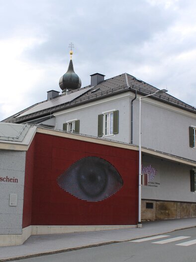 Ein modernes Gebäude mit einem auffälligen Wandgemälde eines Auges. Die Umgebung ist urban, mit einem ruhigen Straßenabschnitt. | © TV Südsteiermark - Irene Löschnig