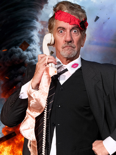 A man in a suit is holding a phone and looks surprised. In the background, a destructive tornado is visible. | © Neue Bühne Wien