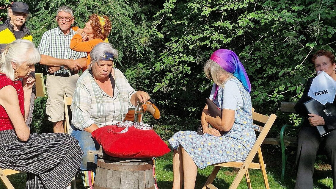 Eine Gruppe von Personen sitzt im Freien und genießt ihre Zeit zusammen. Einige Frauen unterhalten sich und bereiten etwas auf einem Tisch vor. | © TRUNK-Elfi Greiner