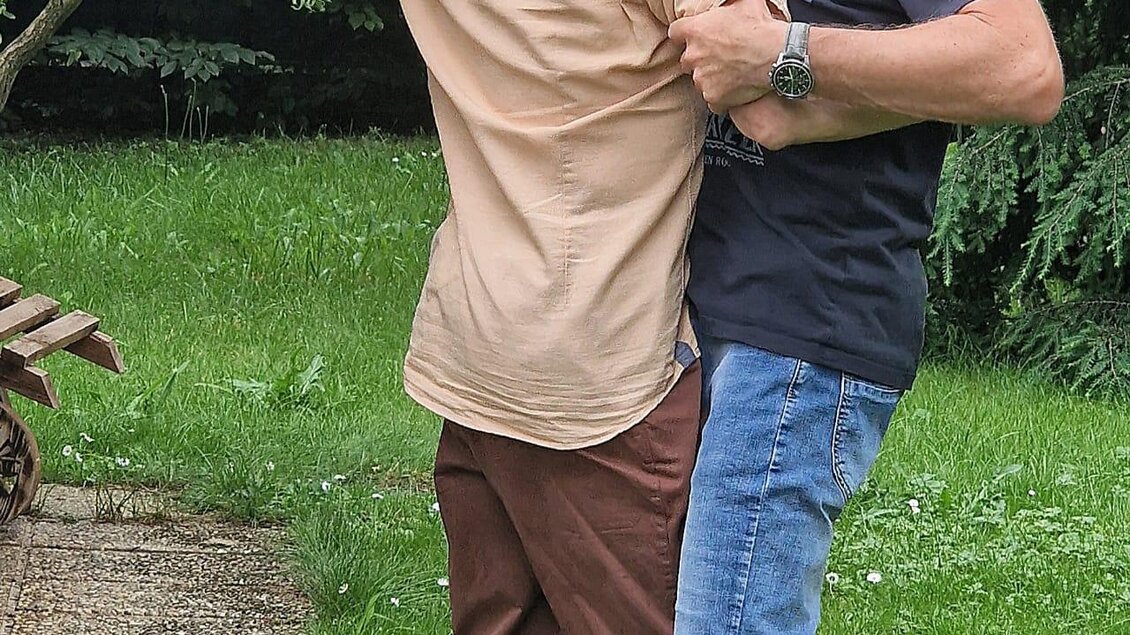 Zwei Männer stehen in einem Garten und umarmen sich in einer spielerischen Pose. Im Hintergrund sind Bäume und grünes Gras zu sehen. | © TRUNK-Elfi Greiner
