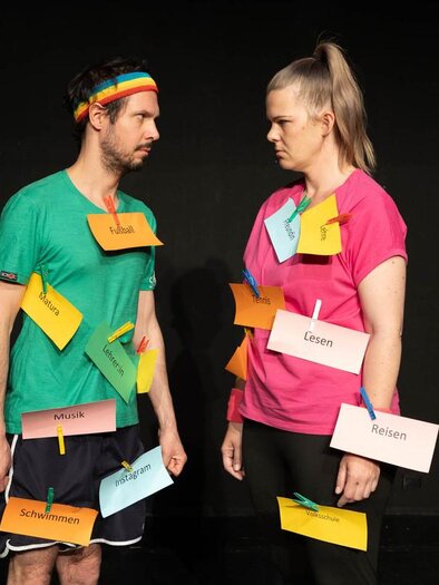 Zwei Personen stehen sich gegenüber, beide tragen bunte T-Shirts und haben Kärtchen um ihren Hals. Die Kärtchen zeigen verschiedene Wörter und Begriffe, die möglicherweise eine Diskussion oder ein Spiel darstellen. | © Christian Koschar