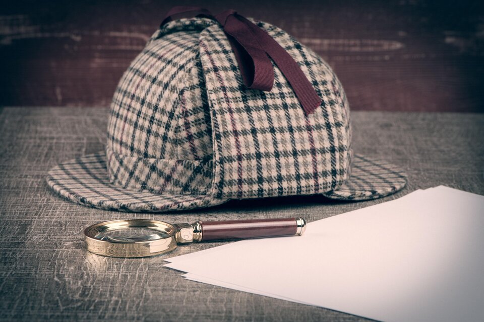 Karrierter Hut von Sherlock Holmes mit Lupe und Papierblätter auf einem Tisch.  | © AdobeStock