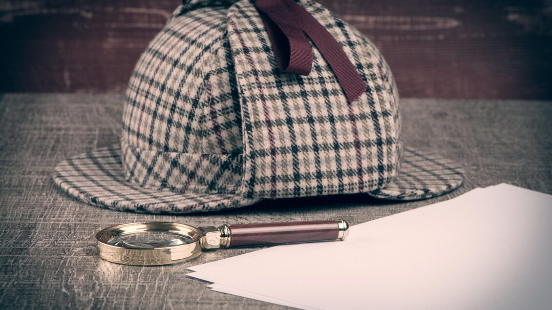 Karrierter Hut von Sherlock Holmes mit Lupe und Papierblätter auf einem Tisch.  | © AdobeStock