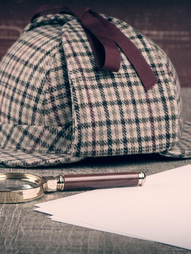Karrierter Hut von Sherlock Holmes mit Lupe und Papierblätter auf einem Tisch.  | © AdobeStock