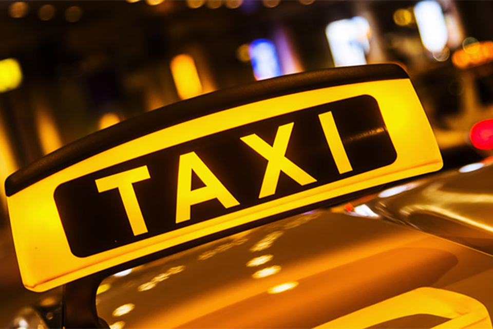 Ein leuchtendes Taxi-Schild mit der Aufschrift "TAXI". Es befindet sich im nächtlichen Stadtbereich mit verschwommenen Lichtern im Hintergrund. | © Komoedie Graz