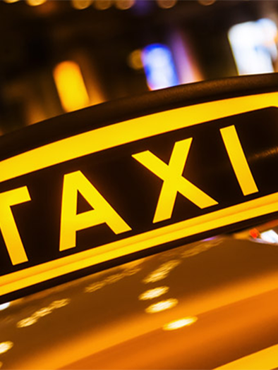A luminous taxi sign with the inscription "TAXI". It is located in the nighttime city area with blurred lights in the background. | © Komoedie Graz