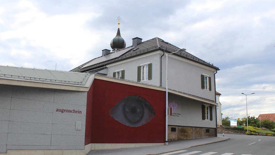 Ein modernes Gebäude mit einem auffälligen Wandgemälde eines Auges. Die Umgebung ist urban, mit einem ruhigen Straßenabschnitt. | © TV Südsteiermark - Irene Löschnig