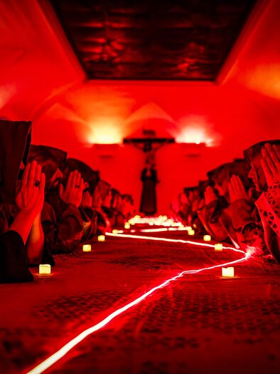 Eine Gruppe von Menschen sitzt in einem schwach beleuchteten Raum mit rotem Licht. Sie beten mit gefalteten Händen vor einer langen, beleuchteten Tischreihe. | © Verein Wandelbühne