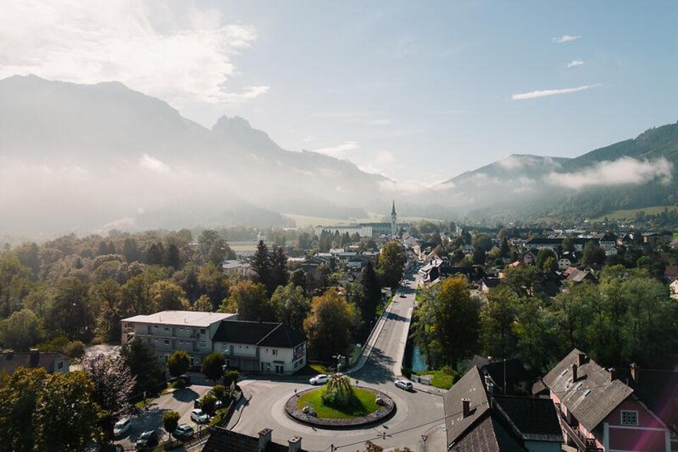 Eine malerische Landschaft mit Bergen und foggy Wolken. Im Vordergrund ist eine ruhige Stadt mit grünen Bäumen und einem Kreisverkehr zu sehen.