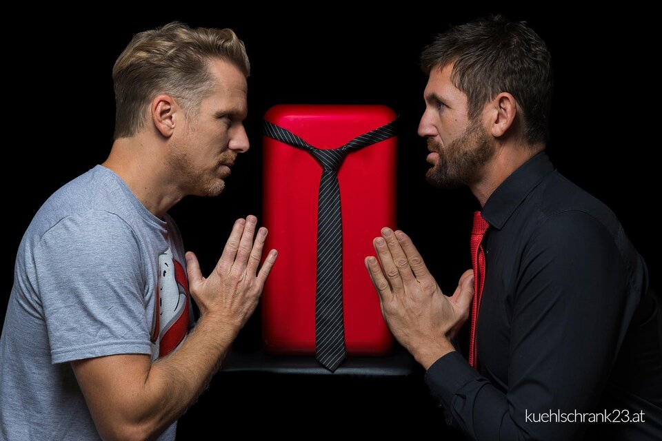 Andreas Ortner und Markus Perner stehen sich auf einer Bühne oder im Studio gegenüber, zwischen ihnen ein roter Kühlschrank mit Krawatte – humorvolle Inszenierung des Kabarett- und Comedy-Duos Kühlschrank23, kreative Unterhaltung und kulturelle Vielfalt in der Stadthalle Fürstenfeld. | © Richard Weiß