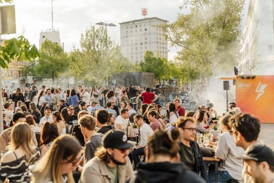 Eine lebhafte Veranstaltung im Freien mit vielen Menschen, die an Tischen sitzen und sich unterhalten. Im Hintergrund sind Bäume und moderne Gebäude sichtbar. | © lendstrom