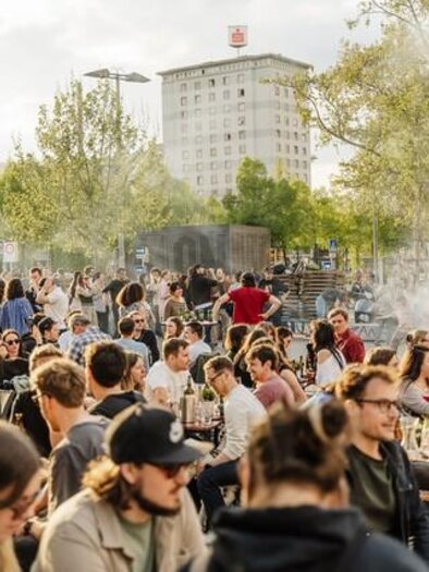 Eine lebhafte Veranstaltung im Freien mit vielen Menschen, die an Tischen sitzen und sich unterhalten. Im Hintergrund sind Bäume und moderne Gebäude sichtbar. | © lendstrom