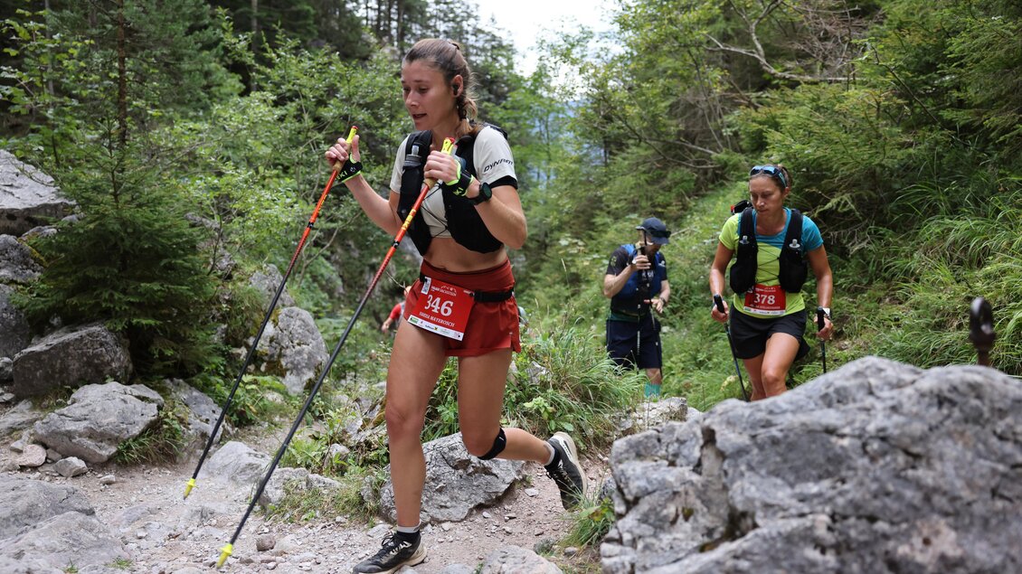 Eine Gruppe von Sportlern läuft einen steilen Wanderweg in einer bewaldeten Umgebung. Die Teilnehmer nutzen Trekkingstöcke und tragen sportliche Kleidung. | © Torlauf Dachstein 2026