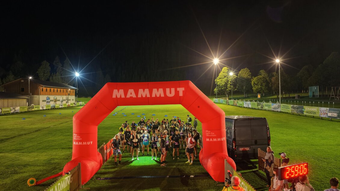 Eine Gruppe von Läufern steht unter einem großen roten Bogen mit dem Schriftzug "MAMMUT". Die Szene spielt sich nachts auf einem Sportplatz ab, mit beleuchteten Bäumen im Hintergrund.