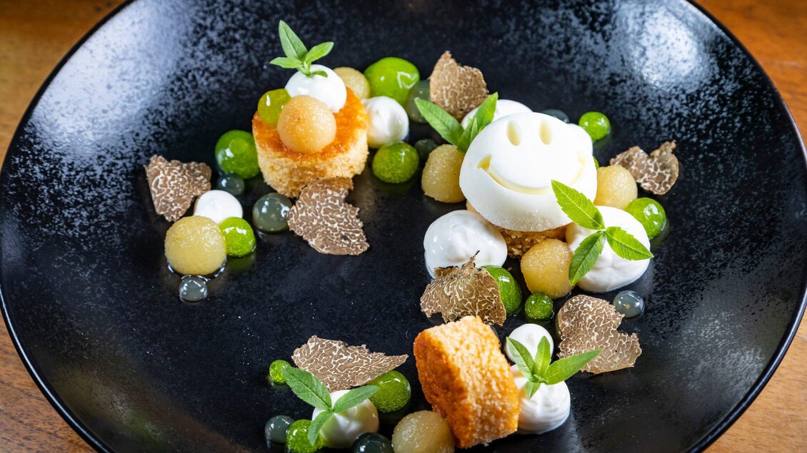 Ein kunstvoll angerichtetes Dessert auf einem schwarzen Teller. Es besteht aus verschiedenen Fruchtkugeln, Schlagsahne und einer lächelnden Eiskugel. | © Graz Tourismus - Werner Krug