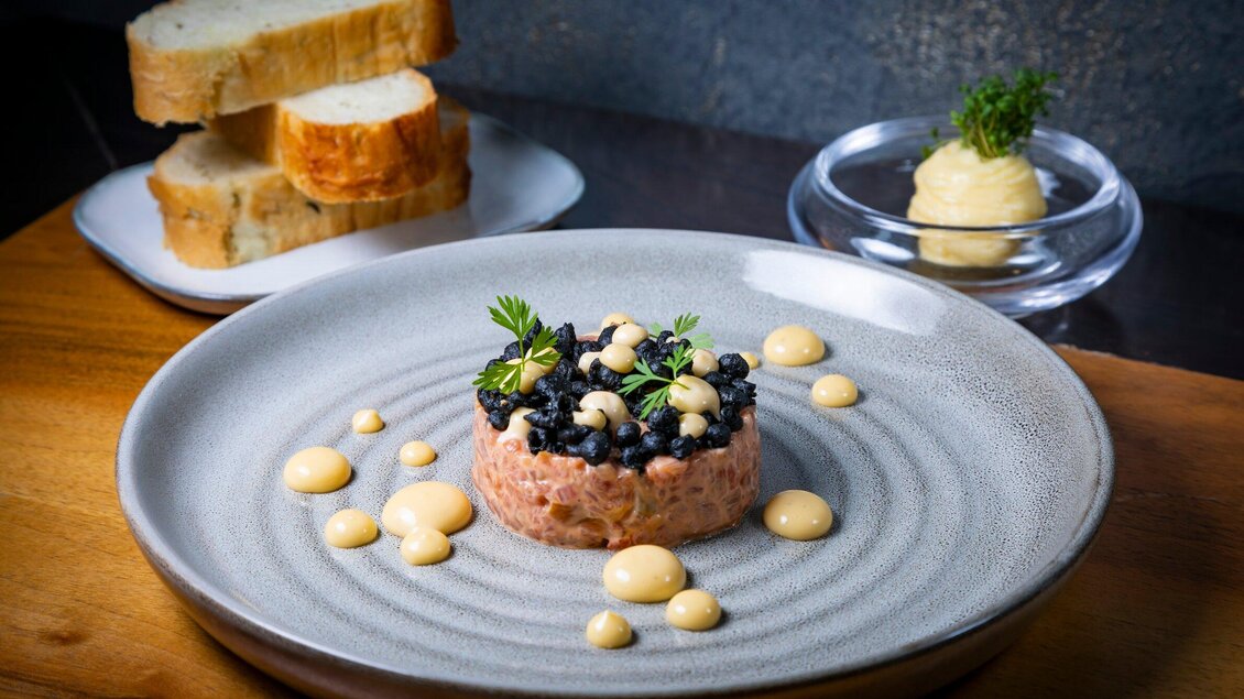 Ein kunstvoll angerichteter Teller mit Lachs-Tatar, garniert mit schwarzen Beeren und einer cremigen Sauce. Im Hintergrund liegen frisches Brot und eine Portion Butter. | © Graz Tourismus - Werner Krug