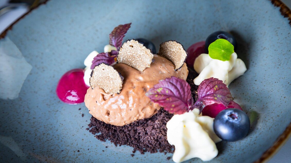 Ein kunstvoll angerichtetes Dessert auf einem blauen Teller. Es besteht aus Schokoladeneis, garnierte mit Trüffelscheiben, Beeren und Sahne. | © Graz Tourismus - Werner Krug