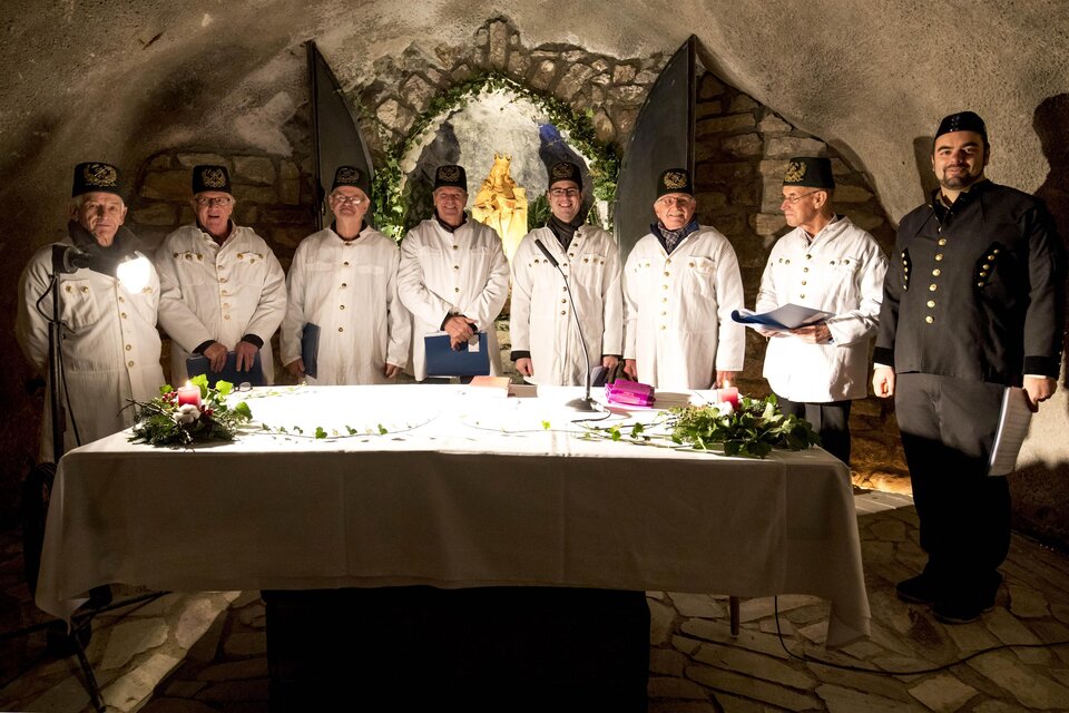 Eine Gruppe von Männern in weißen Gewändern steht um einen Tisch in einer steinernen Höhle. Es gibt Blumen auf dem Tisch und eine dekorative Kulisse im Hintergrund. | © August Zoebl
