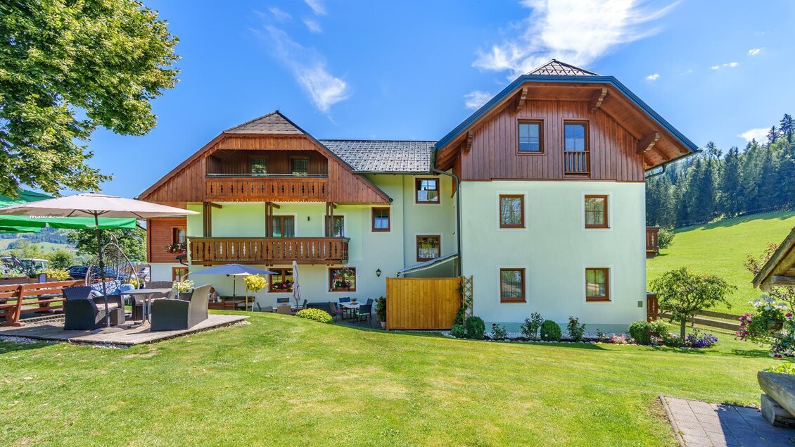 Ein hübsches Haus mit einem Garten und einer Terrasse. Im Hintergrund sind grüne Hügel und ein blauem Himmel zu sehen.