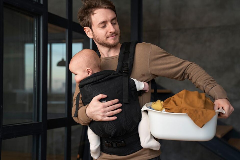 Ein Vater trägt ein Baby in einem Tragetuch und hält mit der anderen Hand einen Korb. Der Raum hat eine moderne und helle Atmosphäre. | © Bild: Freepik