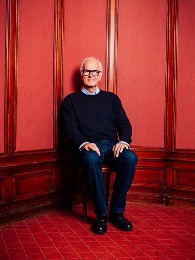 An older man is sitting on a chair in a corner of a room. The walls are red and the decor appears classic.