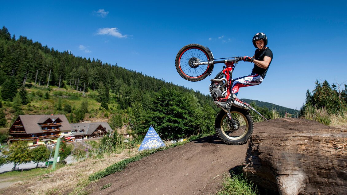 Ein Fahrer macht einen Wheelie auf seinem Motorrad auf einem Waldweg. Im Hintergrund sind Bäume und ein paar Gebäude sichtbar. | © Salzstiegl-Tom Lamm