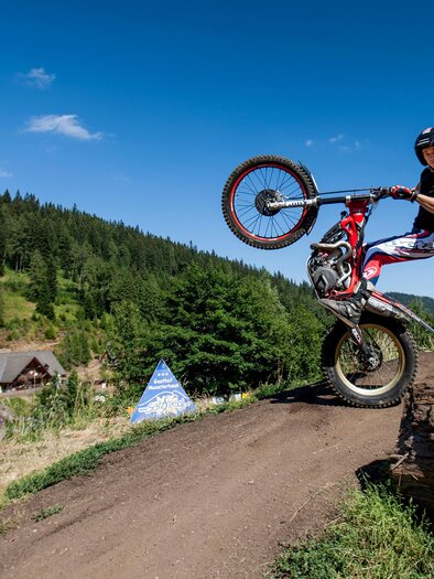 Ein Fahrer macht einen Wheelie auf seinem Motorrad auf einem Waldweg. Im Hintergrund sind Bäume und ein paar Gebäude sichtbar. | © Salzstiegl-Tom Lamm