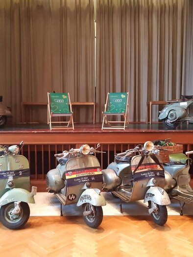 A series of historical Vespas are standing on a stage. In the background, two green chairs and a banner with information about the region of Graz can be seen. | © Trofeo IV Del VC Köflach
