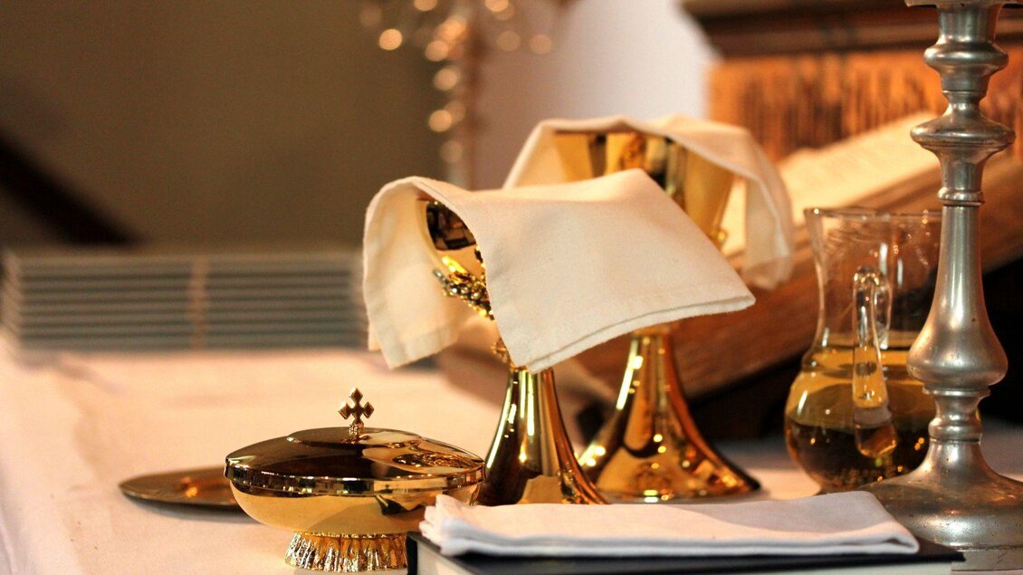 Ein Tisch mit goldenen Kelchen und einem Kreuz. Daneben stehen eine Glasflasche und ein aufgeschlagenes Buch. | © Tieber Doris
