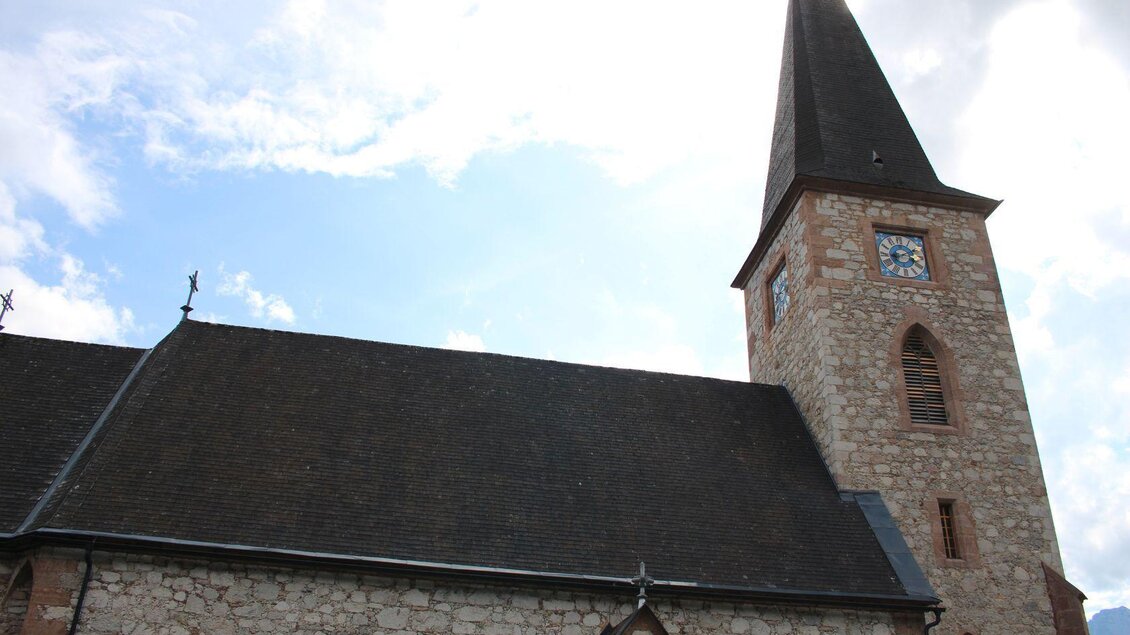 Eine alte Steinkirche mit einem hohen, spitzen Turm. Der Himmel ist teilweise bewölkt. | © Viola Lechner