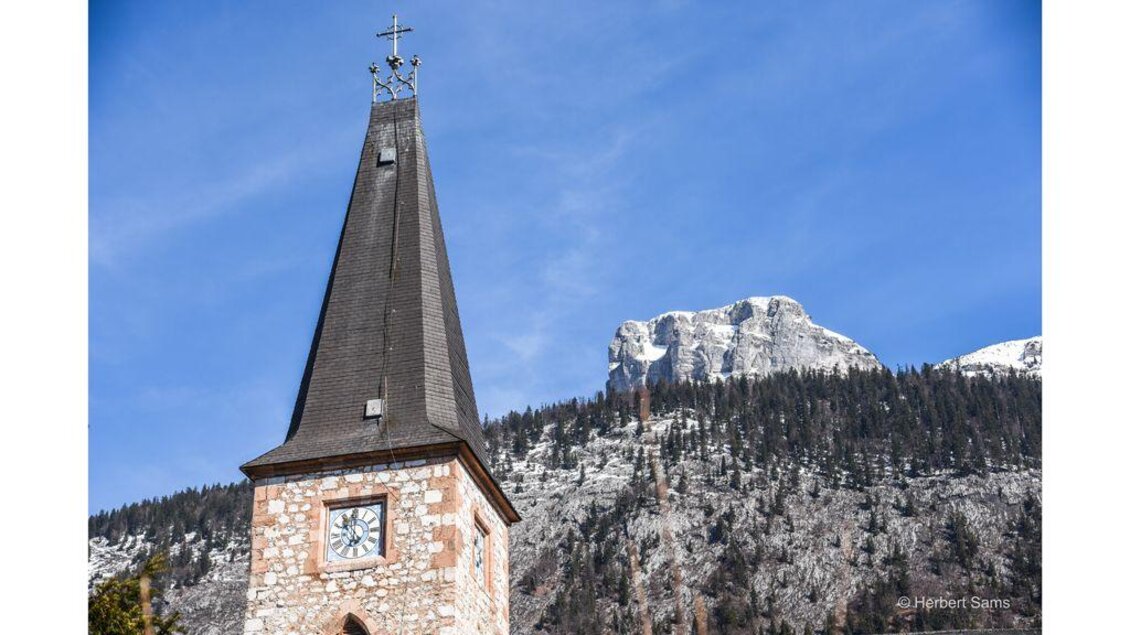 Ein Kirchturm mit einem charakteristischen Spitzdach und einer Uhr. Im Hintergrund sind schneebedeckte Berge und bewaldete Hänge zu sehen. | © Herbert Sams