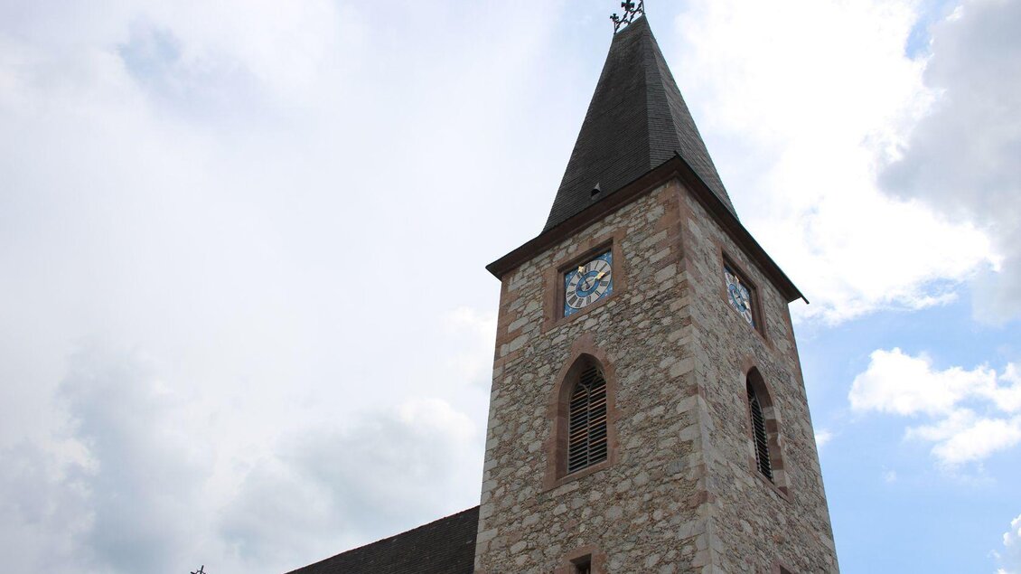 Eine steinerne Kirchturm mit einem spitzen Dach und einem Uhrenblatt. Der Himmel ist bewölkt und es ist tagsüber. | © Viola Lechner