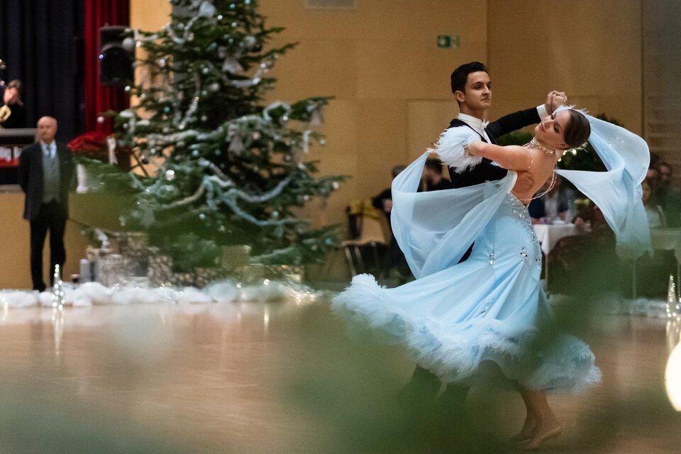 Ein Paar tanzt elegant in einem festlich geschmückten Raum. Im Hintergrund steht ein Weihnachtsbaum und Musiker spielen. | © Dance Unity