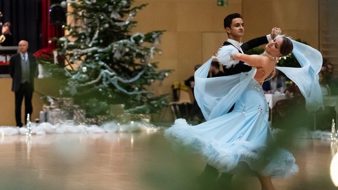 Ein Paar tanzt elegant in einem festlich geschmückten Raum. Im Hintergrund steht ein Weihnachtsbaum und Musiker spielen. | © Dance Unity