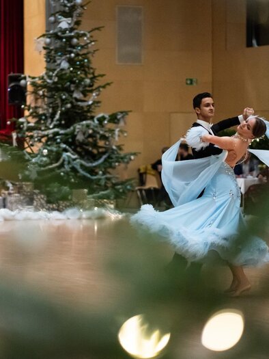 A couple dances elegantly in a festively decorated room. In the background, there is a Christmas tree and musicians are playing. | © Dance Unity