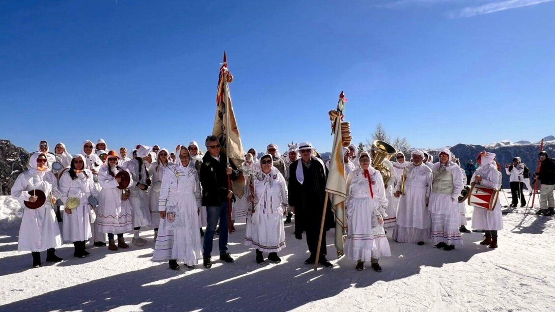 Die Gruppe der Altausseer Trommelweiber in Aufstellung zu ihrem jährlichen Umzug während des Ausseer Faschings. Sie tragen die traditionelle Verkleidung, bestehend aus weißen Nachtgewändern, Schlafhauben und Masken. Sie stehen auf schneebedecktem Boden in Altaussee unter einem strahlend blauen Himmel, und halten Fahnen, Trommeln und Trompeten in ihren Händen. | © Martina Gaisberger