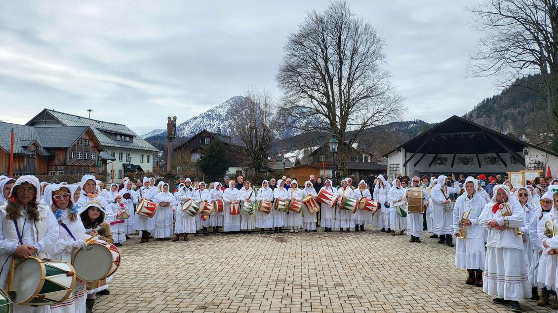 Die Gruppe der Altausseer Trommelweiber in Aufstellung zu ihrem jährlichen Umzug während des Ausseer Faschings. Sie tragen die traditionelle Verkleidung, bestehend aus weißen Nachtgewändern, Schlafhauben und Masken. Sie stehen auf einem Platz in Altaussee unter wolkenbedecktem Himmel, und halten Trommeln und Trompeten in ihren Händen. Im Hintergrund sind die Berge des Ausseerlandes und traditionelle "ausseerische" Gebäude sichtbar. | ©  Martina Gaisberger
