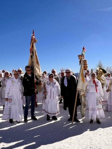 The group of Altaussee "Trommelweiber" drum women lined up for their annual parade during the Ausseer Carnival. They wear traditional costumes consisting of white nightgowns, nightcaps, and masks. They stand on snow-covered ground in Altaussee under a radiant blue sky, holding flags, drums, and trumpets in their hands. | © Martina Gaisberger