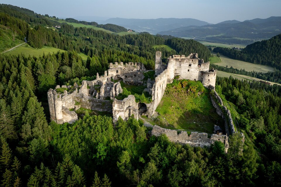 Eine alte Burgruine umgeben von dichten Wäldern und sanften Hügeln. Die Landschaft ist grün und malerisch. | © TV Murau