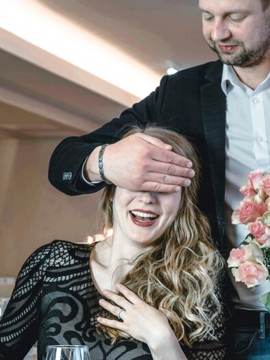 Ein Mann hält die Augen einer Frau mit seiner Hand zu, während sie lächelt. Er hält einen Strauß Rosen in der anderen Hand. | © Vulkanlandhotel Legenstein