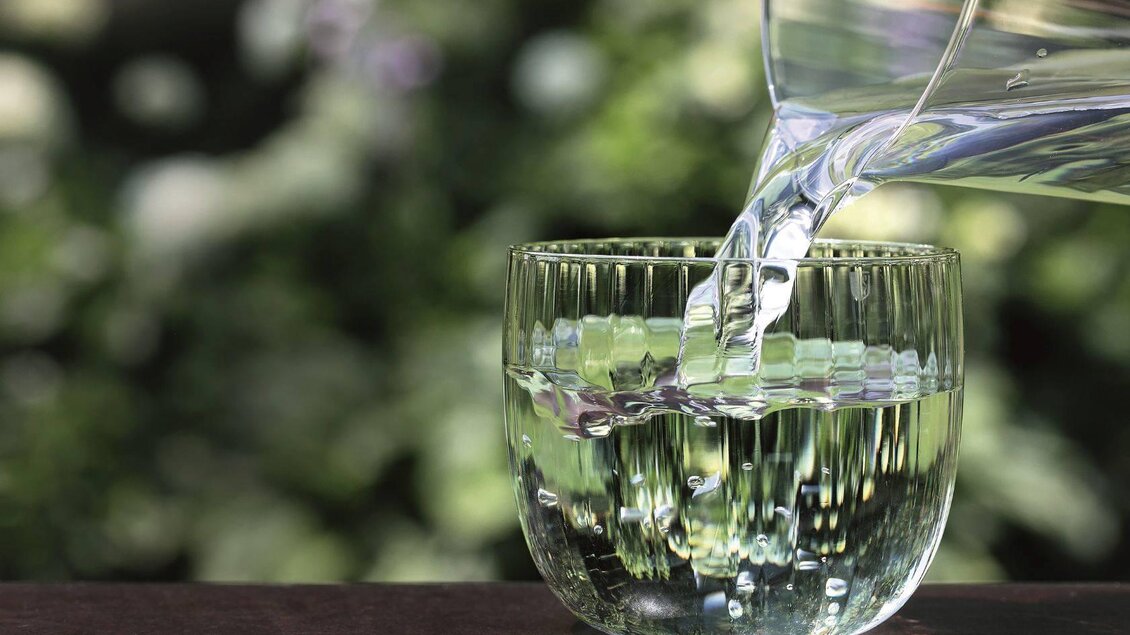 Ein Glas wird mit klarem Wasser gefüllt. Im Hintergrund sind unscharfe grüne Pflanzen zu sehen. | © Stillsegler