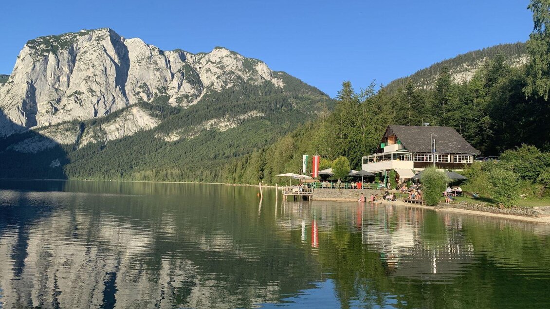 <p>Der ruhige Altausseer See umgeben von der Trisselwand und dichten Wäldern. Im Vordergrund steht das Strandcafe Restaurant Altaussee mit einer Terrasse am Wasser.</p> | © Strandcafe