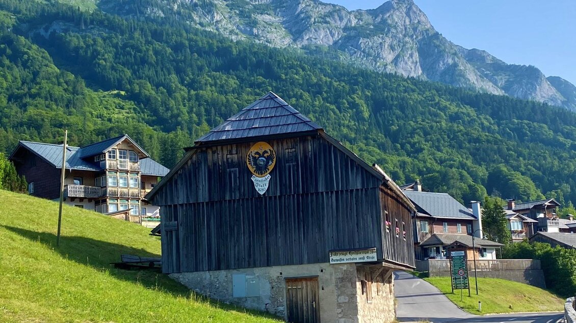Ein traditionelles Holzhaus mit einem Wappen, umgeben von grünen Wiesen und Bergen im Hintergrund. Die klare blaugrüne Landschaft strahlt Ruhe und Idylle aus. | © Bettina Scheck