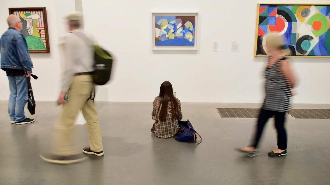 Eine Person sitzt auf dem Boden in einer Kunstgalerie und betrachtet die Ausstellungen. Im Hintergrund sind andere Besucher zu sehen, die an den Gemälden vorbeigehen. | © pixabay