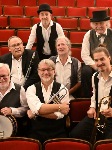 Eine Gruppe von Musikern steht auf einer Bühne in einem Theater. Sie tragen traditionelle Kleidung und halten Musikinstrumente.