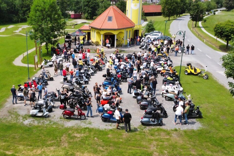 Eine große Gruppe von Menschen versammelt sich mit ihren Vespas am Parkplatz einer Kapelle in der Lipizzanerheimat. Zahlreiche Roller sind aufgereiht, während die Teilnehmer in geselliger Runde zusammenstehen. Im Hintergrund ist die gelbe Kapelle und eine grüne Landschaft zu sehen. | © Uwe Pongratz