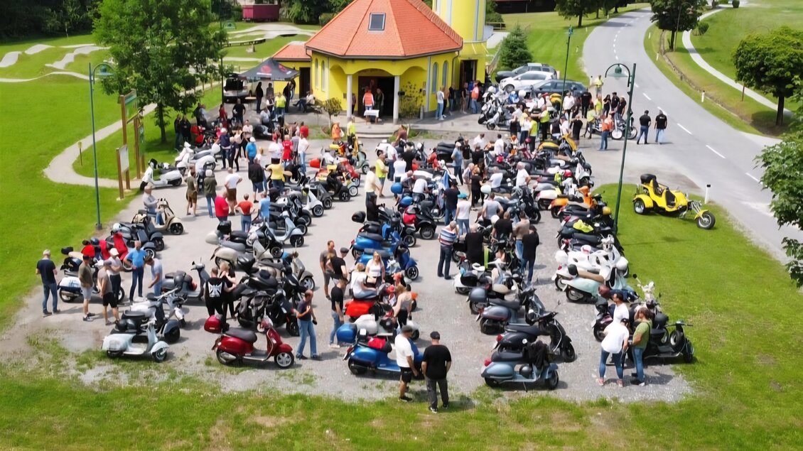 Eine große Gruppe von Menschen versammelt sich mit ihren Vespas am Parkplatz einer Kapelle in der Lipizzanerheimat. Zahlreiche Roller sind aufgereiht, während die Teilnehmer in geselliger Runde zusammenstehen. Im Hintergrund ist die gelbe Kapelle und eine grüne Landschaft zu sehen. | © Uwe Pongratz