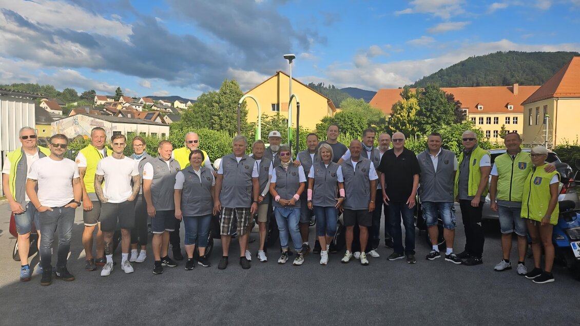 Eine Gruppe von 20 Personen steht in einem sonnigen Außenbereich, umgeben von grünen Hügeln und Häusern. Die Mitglieder des Organisationsteams der Vespaausfahrt in Köflach tragen graue Westen mit Logo und einheitliche Kleidung. Einige tragen auch gelbe Sicherheitswesten. Sie lächeln und posieren für das Foto, während der Himmel teils bewölkt ist. | © Uwe Pongratz